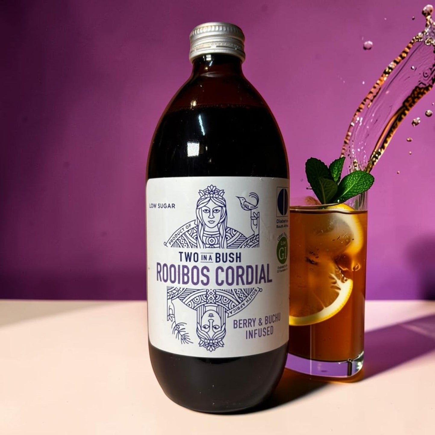 Die Spens Clarens Two In A Bush Berry & Buchu infused low sugar rooibos cordial bottle next to a glass of iced tea with lemon and mint.
