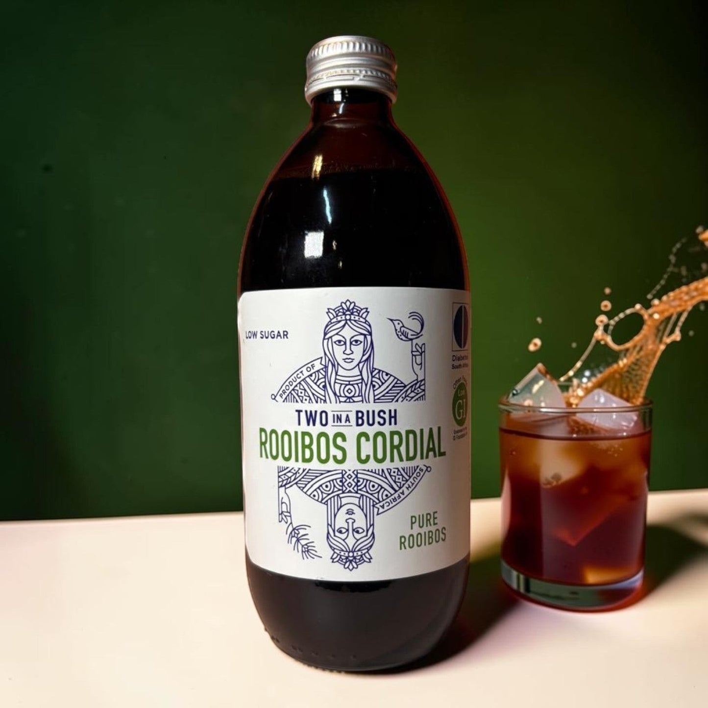 Die Spens Clarens Two In A Bush Pure Rooibos Cordial low sugar in a dark glass bottle next to a glass of iced rooibos cordial.