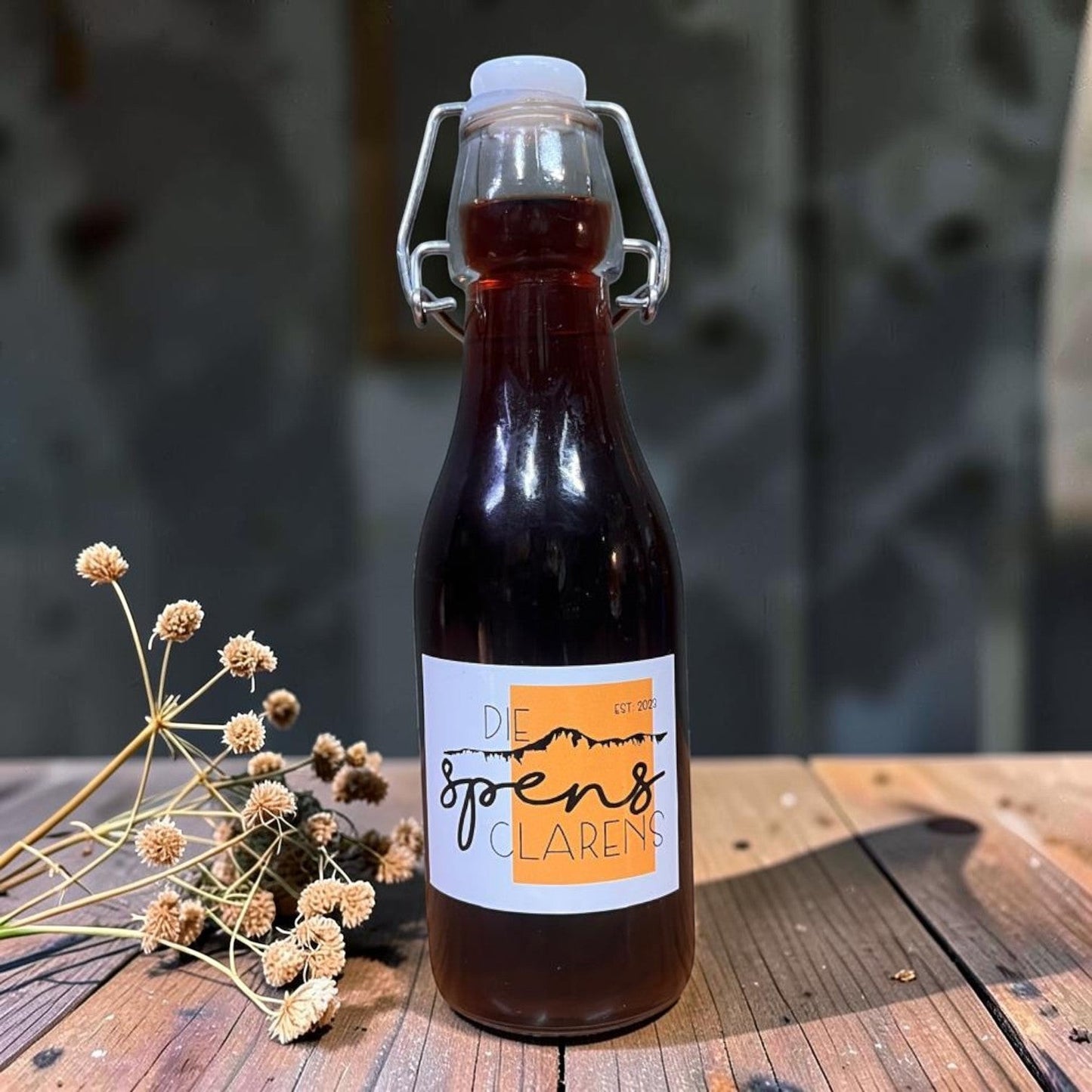 Die Spens Clarens cherry liqueur in a clear glass swing-top bottle, deep ruby hue, on a wooden table with dried flowers.