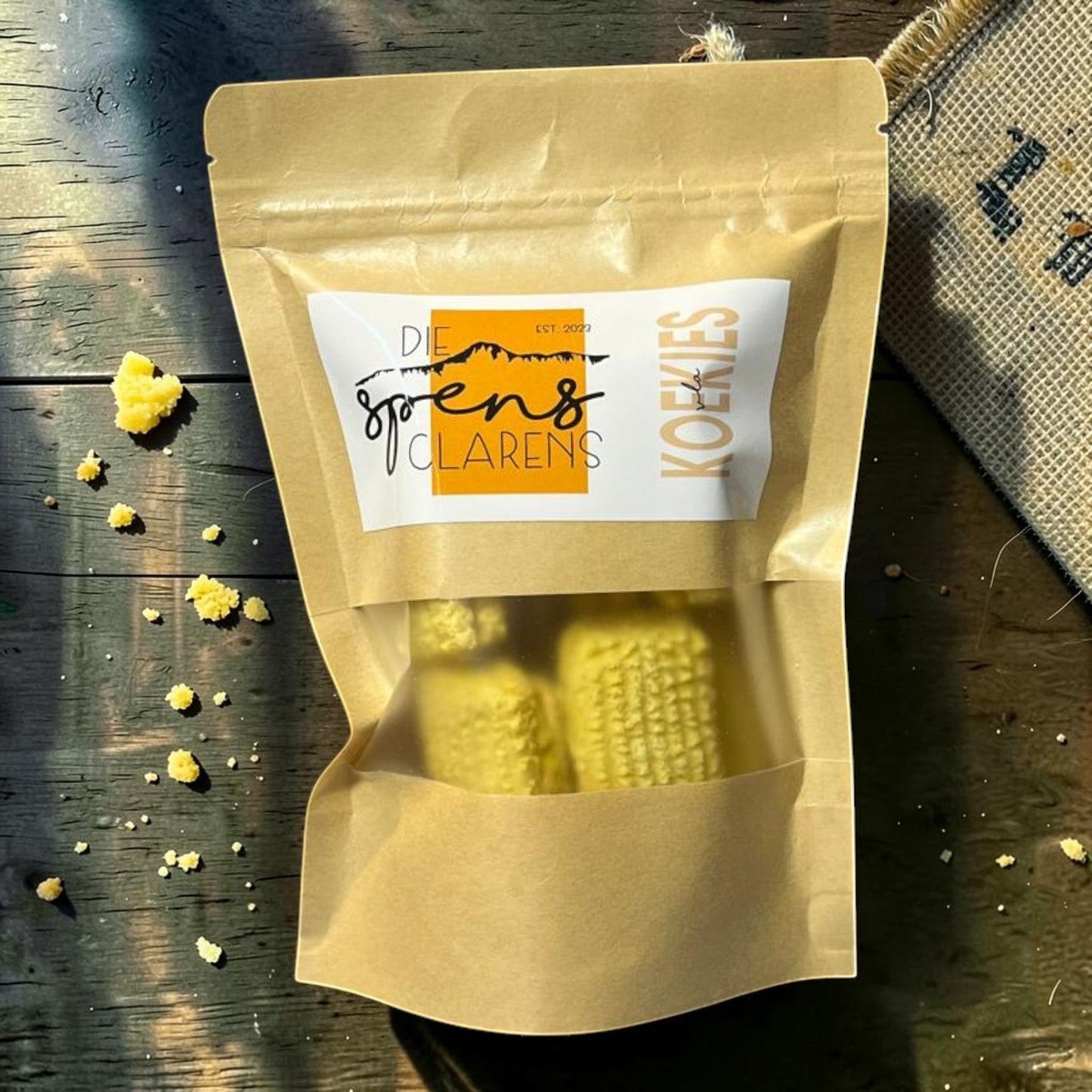 Die Spens Clarens small yellow vla koekies (custard biscuits) in a brown resealable pouch, with crumbs scattered on a wooden surface.