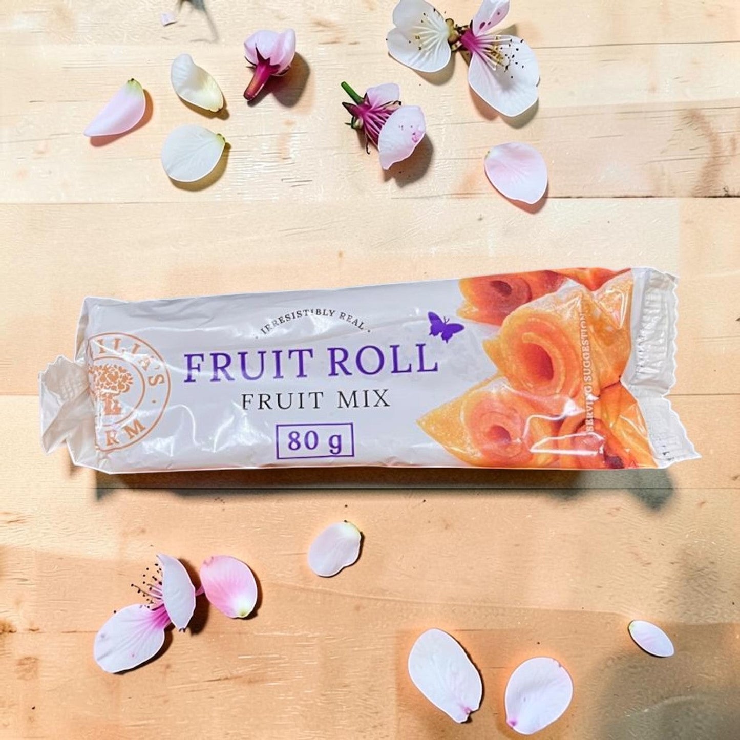 Die Spens Clarens Fruit Roll Fruit Mix 80g, a chewy fruit snack with a vibrant orange hue, surrounded by delicate pink and white flower petals on a wooden surface.