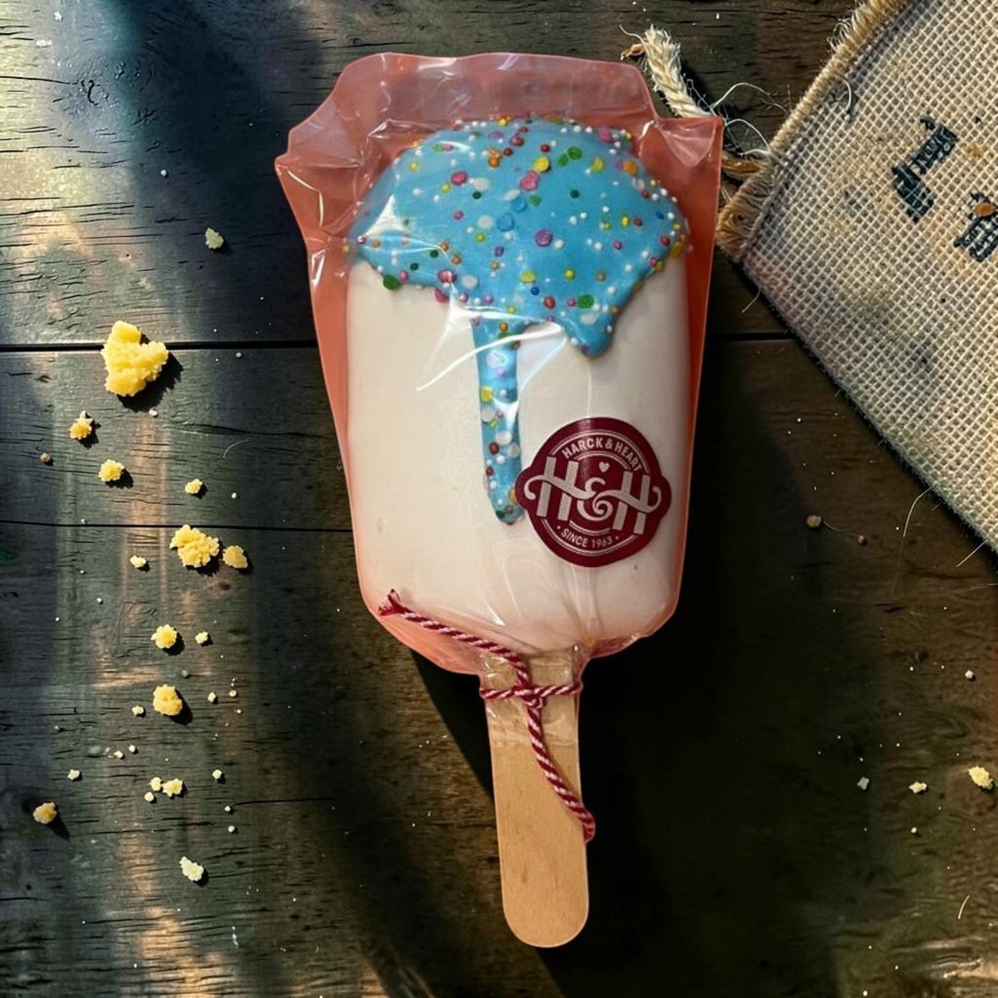 Die Spens Clarens Ice Cream Lolly: white ice cream with blue icing drizzle and colorful sprinkles, on a wooden stick, wrapped in clear plastic.