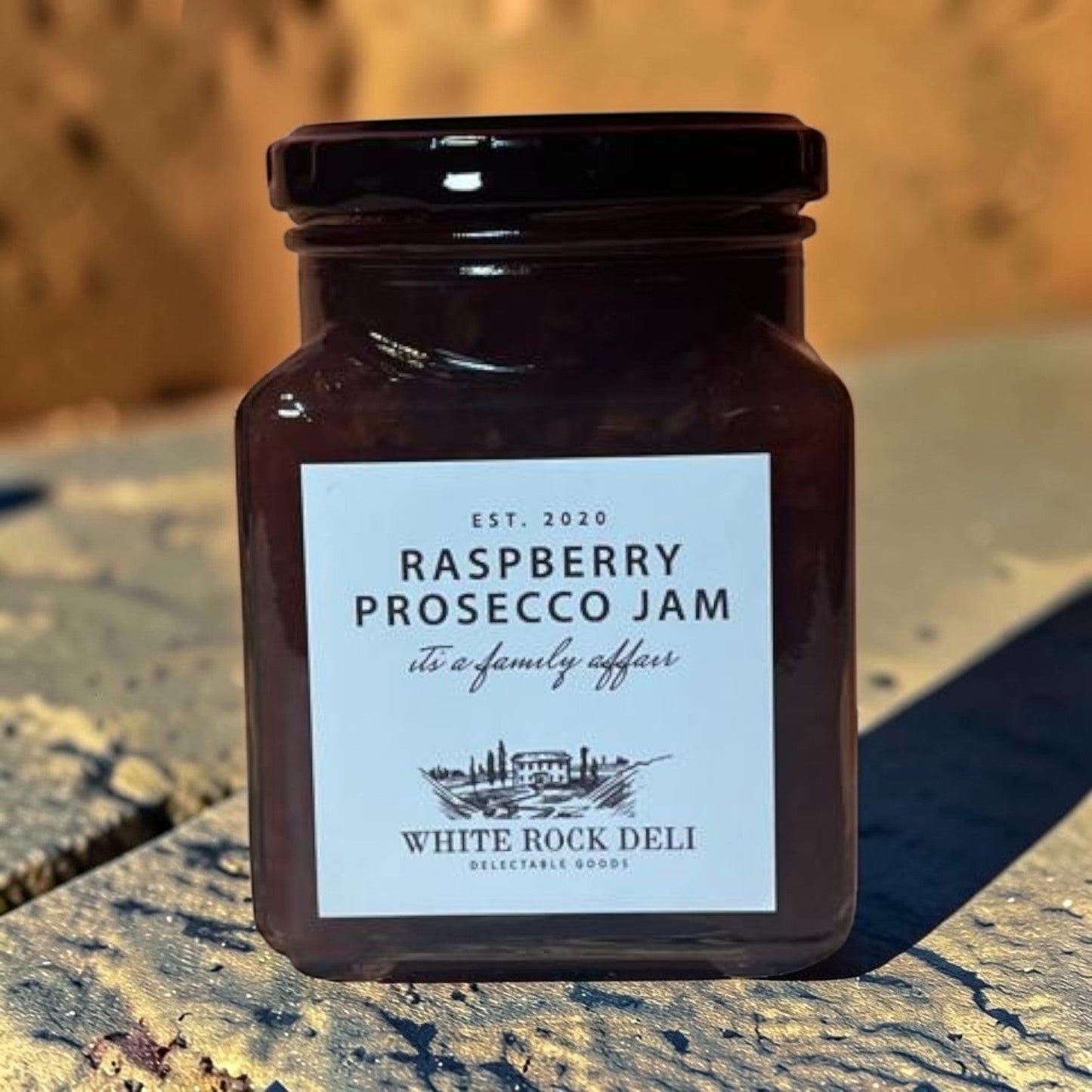 Die Spens Clarens White Rock Deli Raspberry Prosecco Jam in a glass jar with a black lid. The jam is dark red and appears to have fruit pieces.