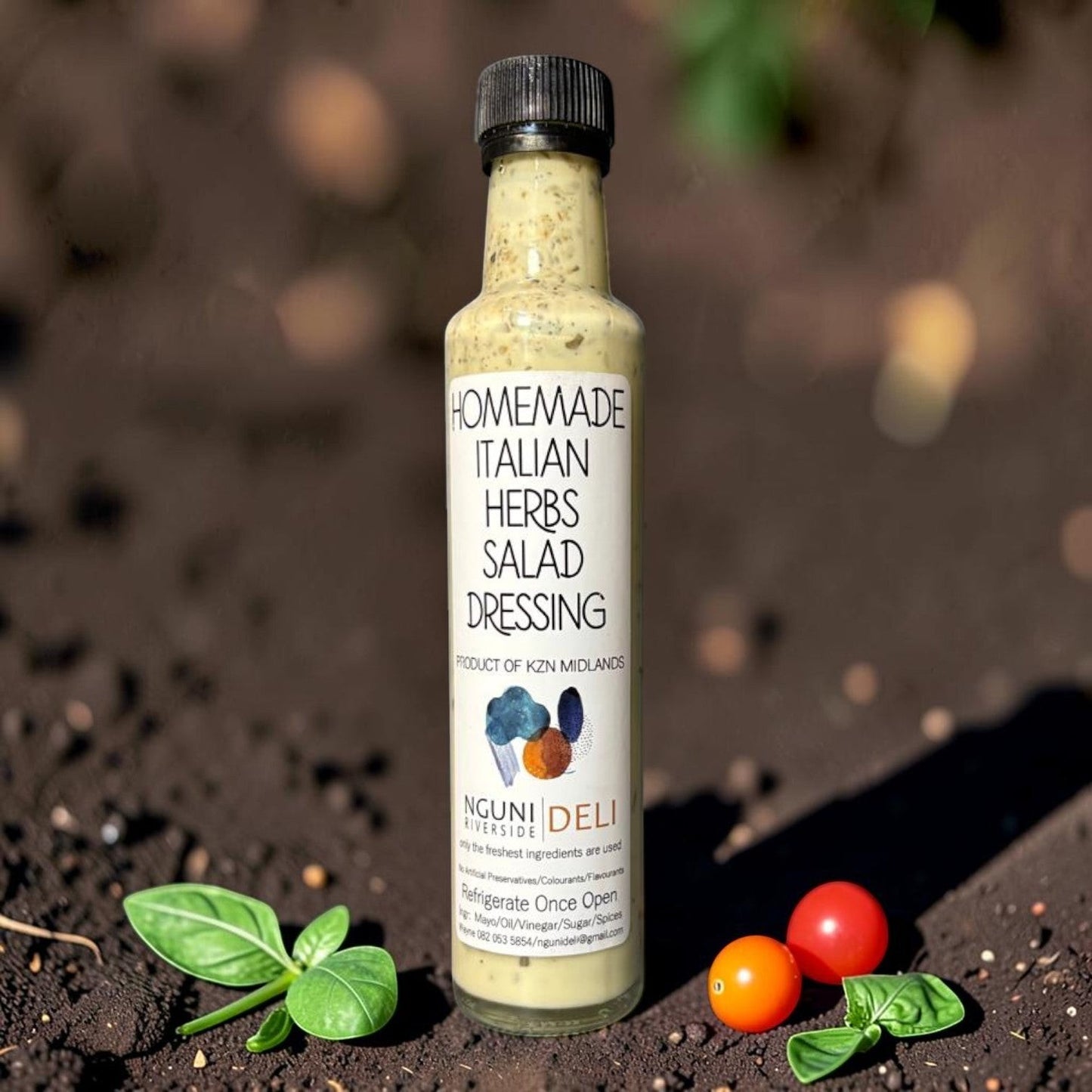 Die Spens Clarens Nguni Deli Italian Herbs Salad Dressing in a glass bottle with a black cap, surrounded by fresh basil and cherry tomatoes on dark soil.