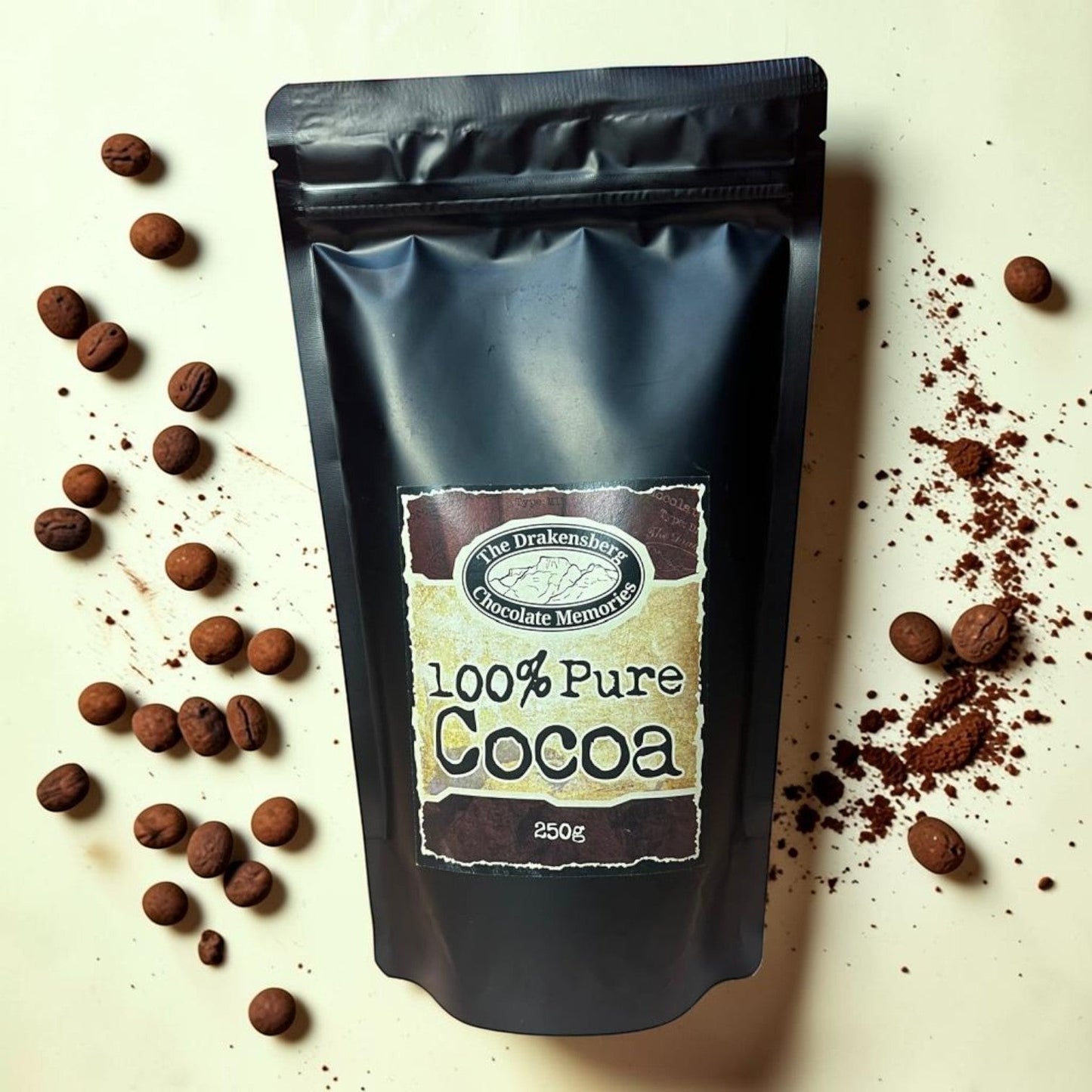 Die Spens Clarens Drakensberg Chocolate Memories 100% Pure Cocoa powder in a black resealable pouch, surrounded by cocoa nibs and powder.