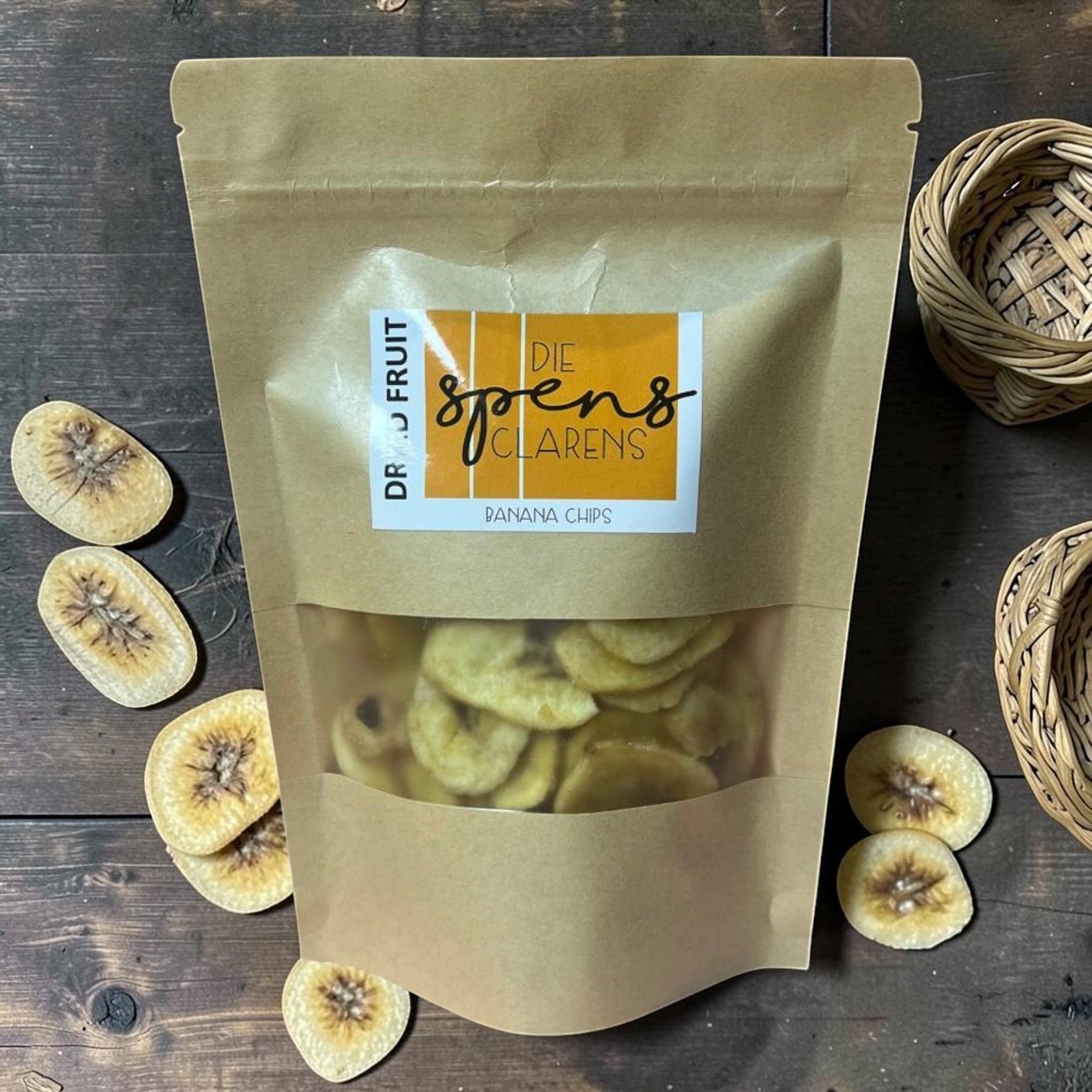 Die Spens Clarens banana chips in a brown resealable bag, surrounded by dried banana slices on a wooden surface.