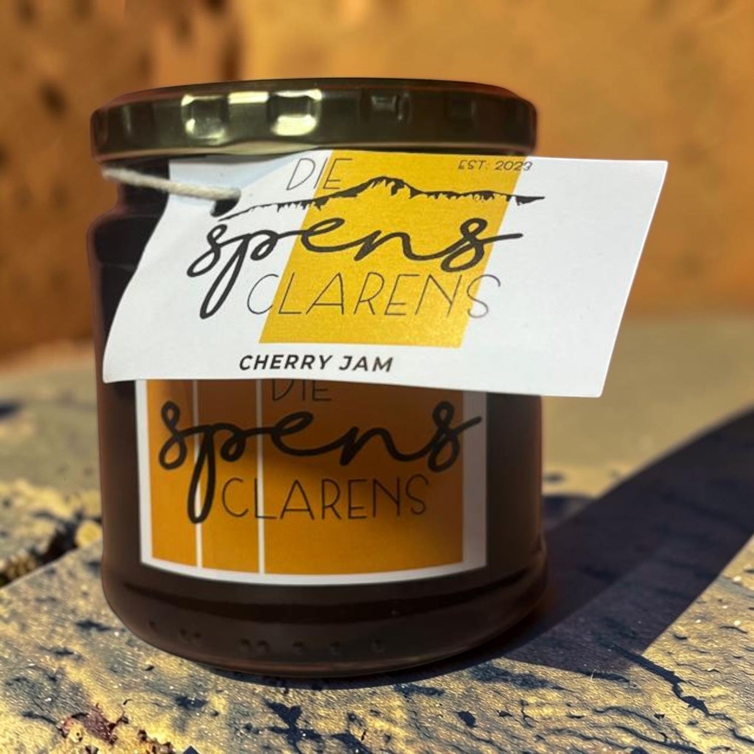 Die Spens Clarens artisanal cherry jam in a glass jar with a gold lid and a white label featuring mountain graphics and brand logo.
