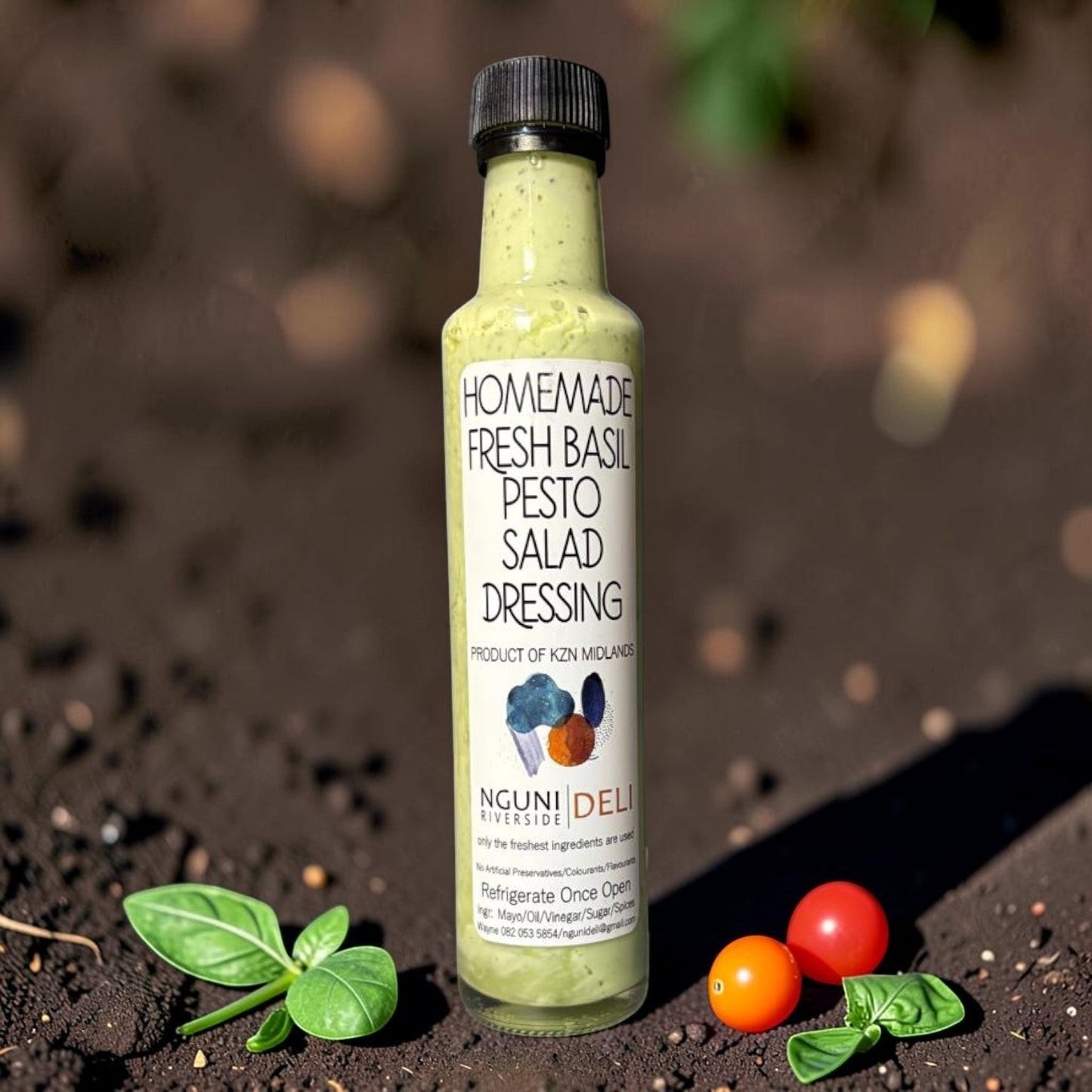 Die Spens Clarens Nguni Deli homemade fresh basil pesto salad dressing in a glass bottle with a black cap, surrounded by fresh basil leaves and cherry tomatoes.