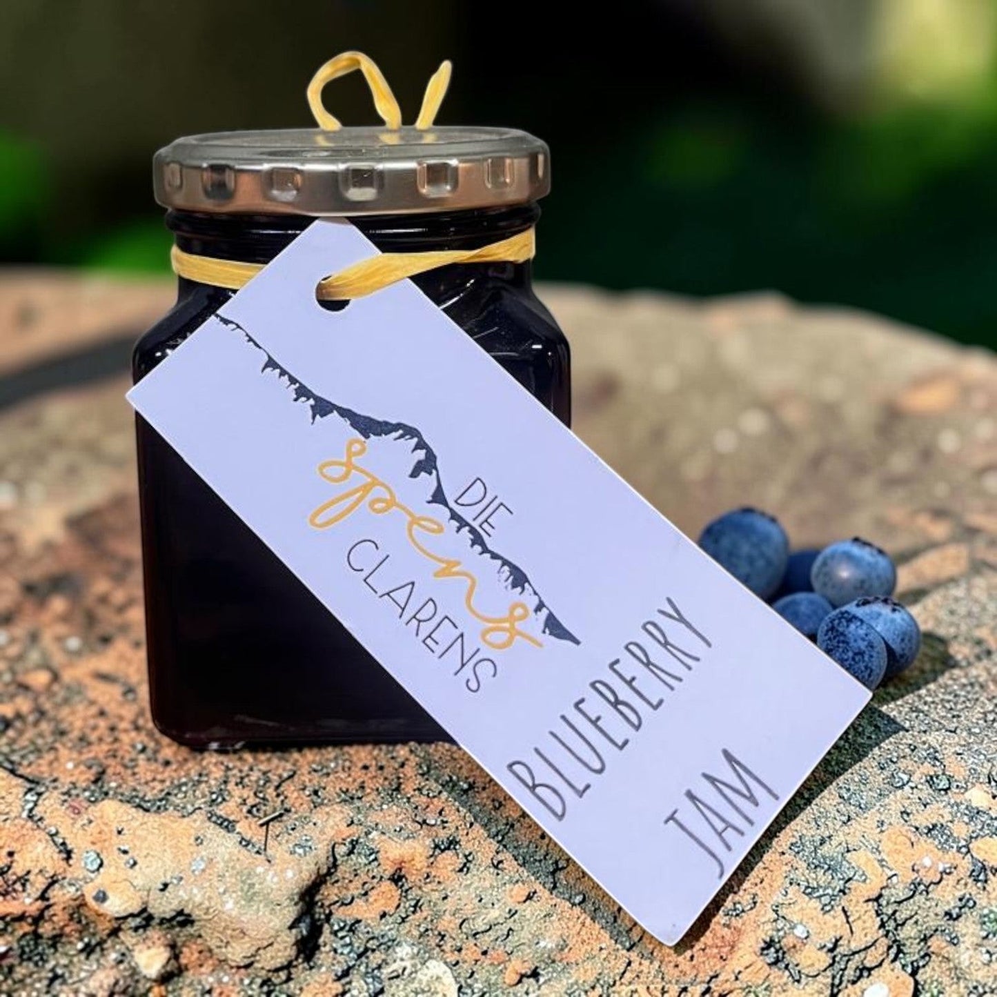 Die Spens Clarens artisanal blueberry jam in a glass jar with a decorative tag and fresh blueberries.