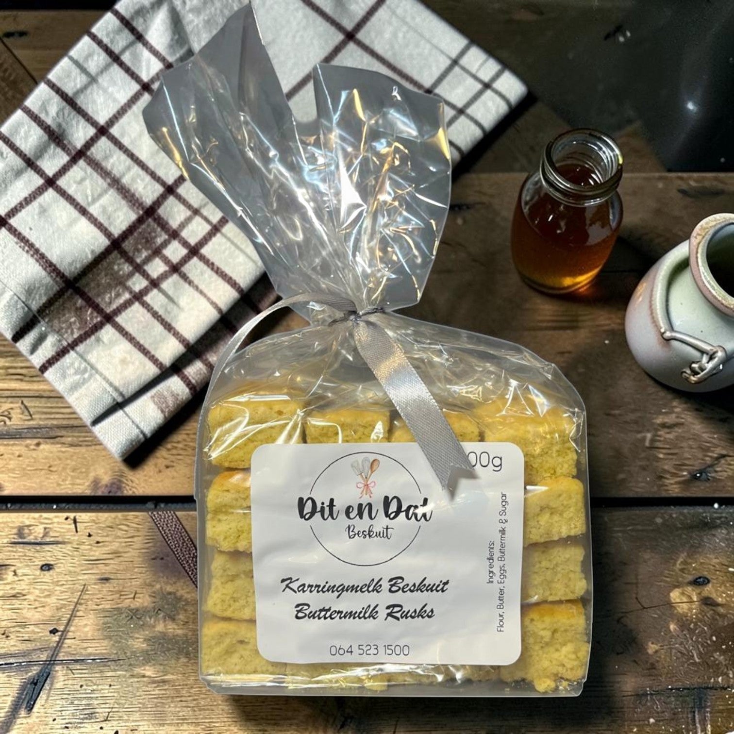 Die Spens Clarens Karringmelk Beskuit (Buttermilk Rusks) in a clear plastic bag tied with a silver ribbon. Golden-brown, square rusks are stacked neatly inside. A label reads 'Dit en Dat Beskuit Karringmelk Beskuit Buttermilk Rusks'.