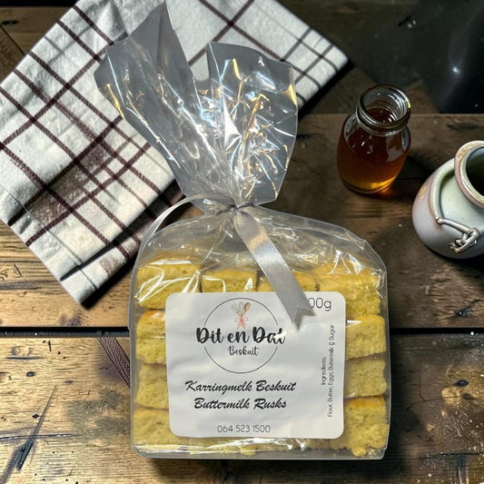 Die Spens Clarens Karringmelk Beskuit (Buttermilk Rusks) in a clear plastic bag tied with a silver ribbon. Golden-brown, square rusks are stacked neatly inside. A label reads 'Dit en Dat Beskuit Karringmelk Beskuit Buttermilk Rusks'.