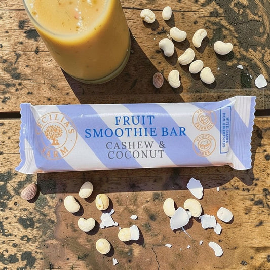 Die Spens Clarens Fruit Smoothie Bar Cashew & Coconut, a vegan and gluten-free snack bar with a blue and white striped wrapper, surrounded by cashews and a glass of smoothie.