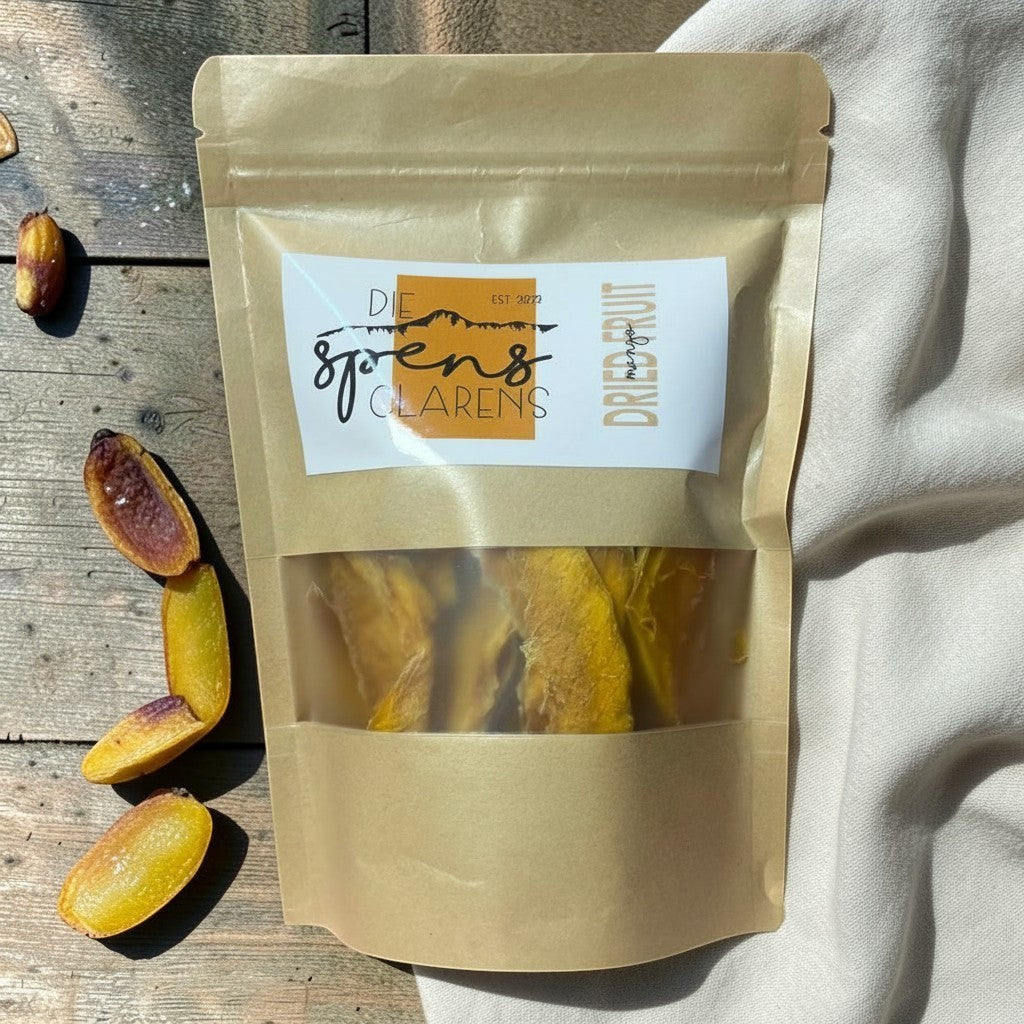 Die Spens Clarens dried mango slices in a resealable pouch with a clear window, showcasing the vibrant yellow fruit. Some dried mango pieces are scattered on a wooden surface.