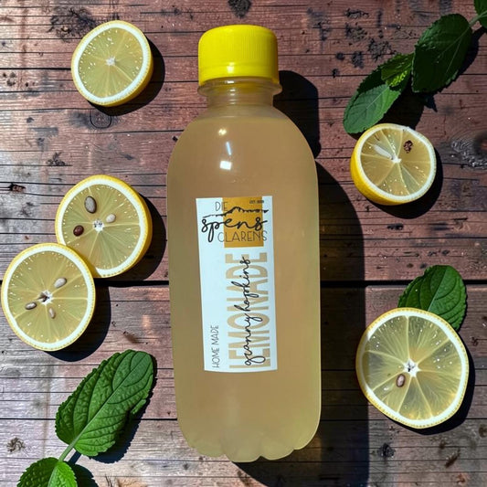Die Spens Clarens Granny Hopkins Lemonade in a clear bottle with a yellow cap, surrounded by lemon slices and mint leaves on a wooden background.