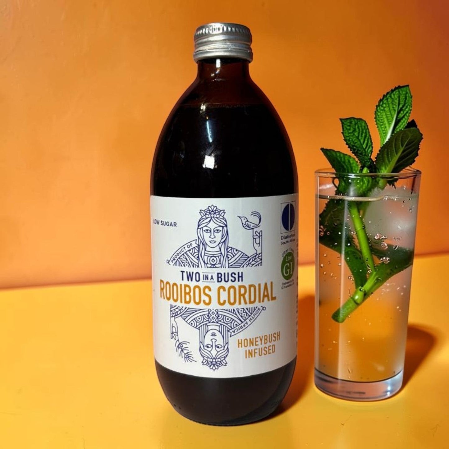 Die Spens Clarens Two In A Bush Rooibos Cordial bottle next to a glass of sparkling water with ice and mint.