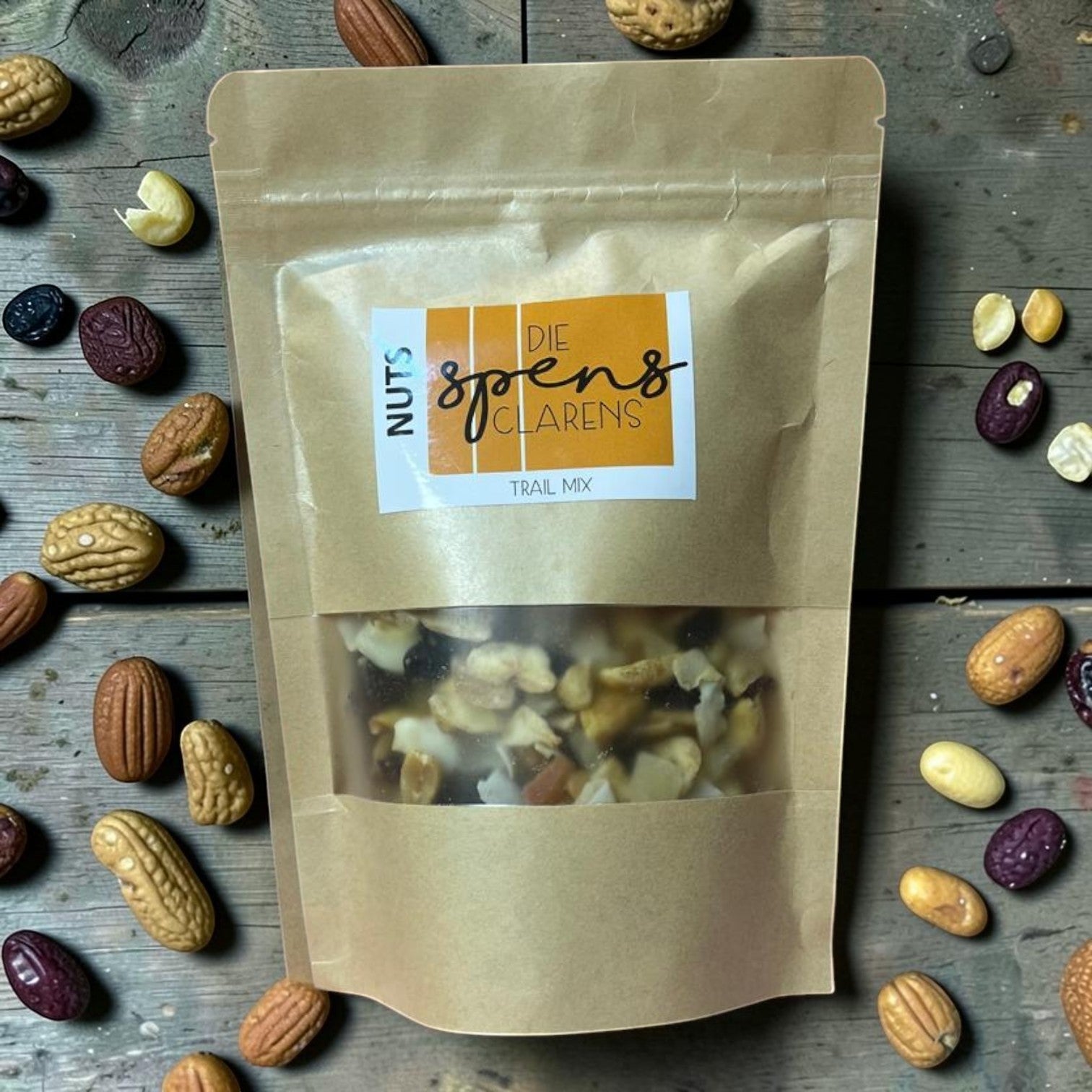 Die Spens Clarens trail mix in a brown paper bag with a clear window showing nuts and dried fruit. Surrounded by loose nuts and dried berries.