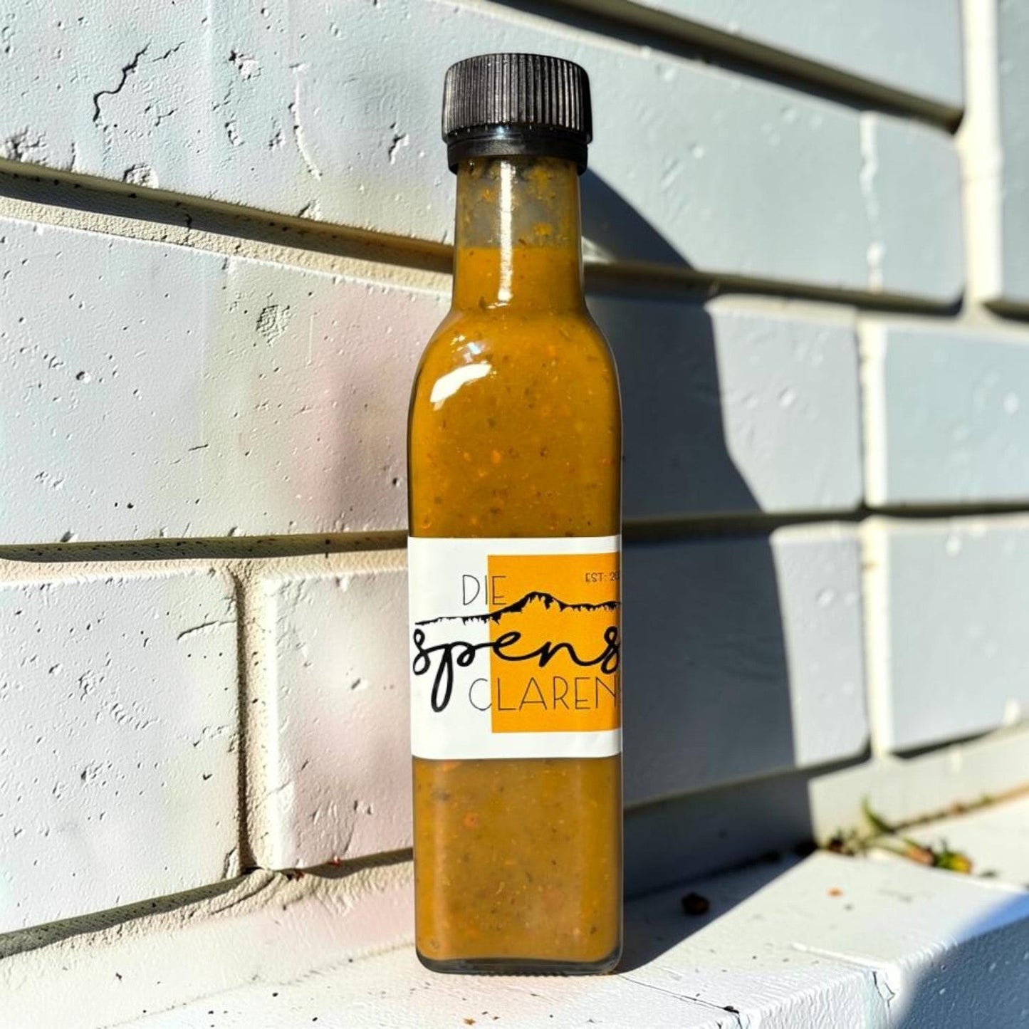 Die Spens AC Hot Sauce bottle with golden hue and textured label, featuring mountains and text. Perfect for adding spice to meals.