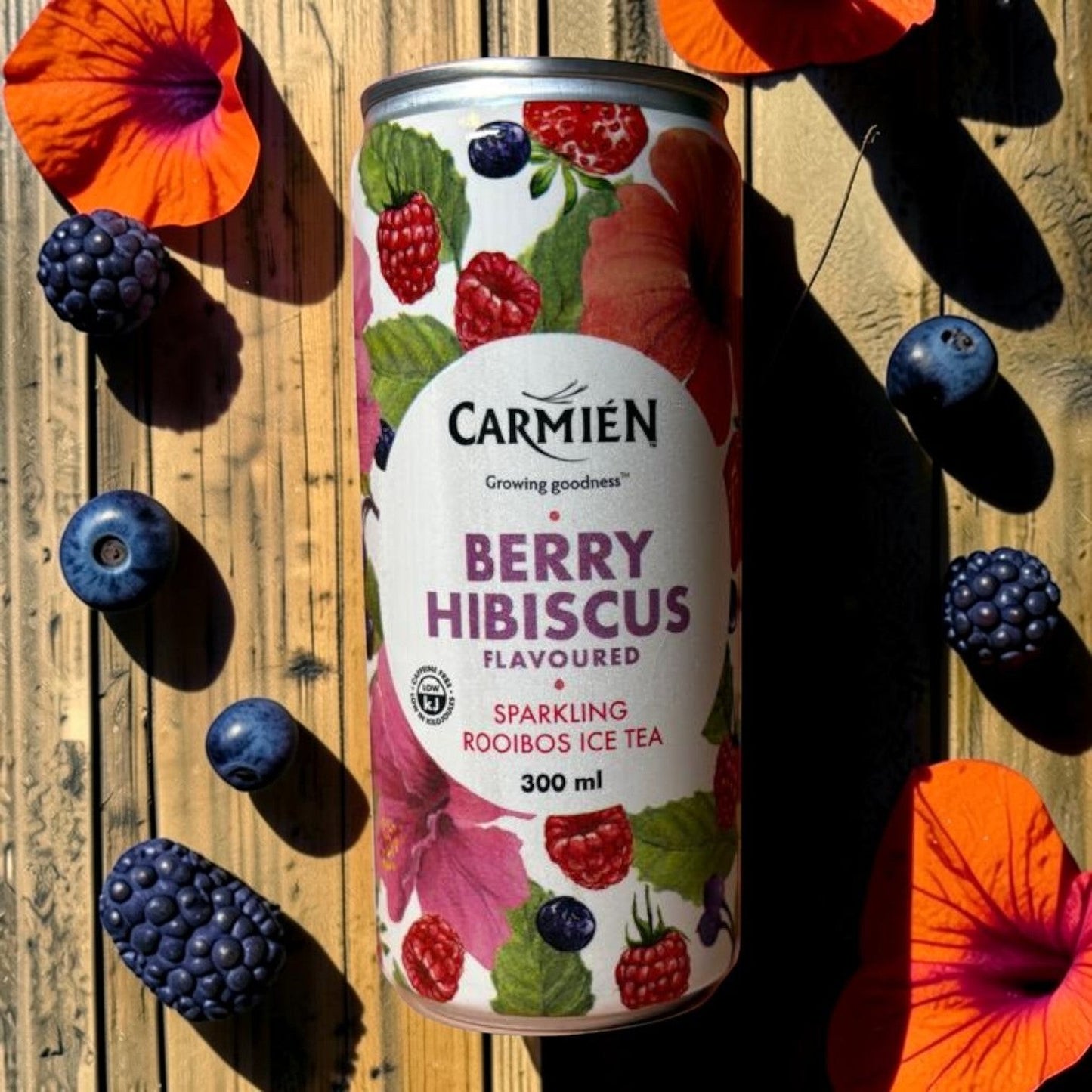 Die Spens Clarens Carmien Berry Hibiscus Sparkling Rooibos Ice Tea can surrounded by fresh berries and hibiscus flowers on a wooden surface.
