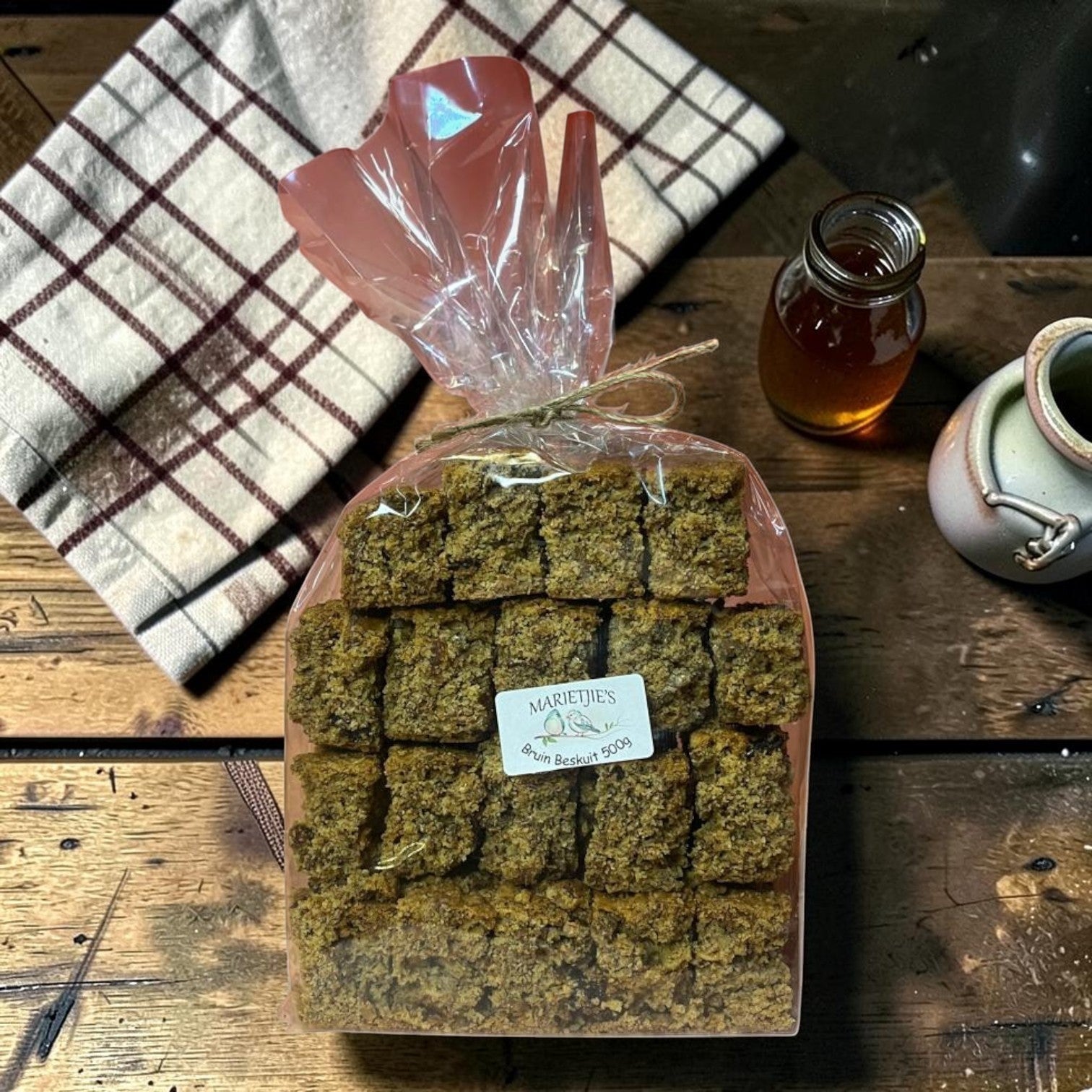 Die Spens Clarens Marietjies Bruin Beskuit rusks in a clear plastic bag, tied with twine. A small bottle of honey and a ceramic jug are visible.