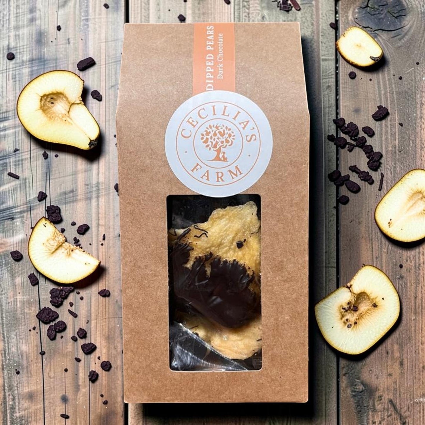 Die Spens Clarens chocolate-dipped peeled pears in a brown box with a window. Sliced pears and dark chocolate pieces surround the box.