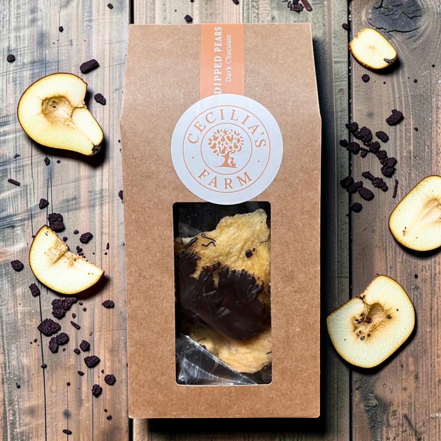 Die Spens Clarens chocolate-dipped peeled pears in a brown box with a window. Sliced pears and dark chocolate pieces surround the box.