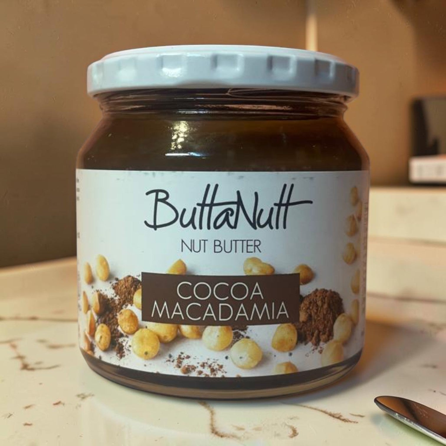 Die Spens Clarens ButtaNutt Cocoa Macadamia nut butter in a glass jar with a white lid, featuring whole macadamia nuts and cocoa powder.