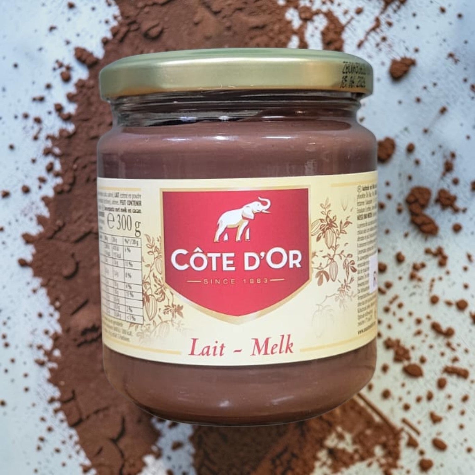 Die Spens Clarens Cote D'Or Lait-Melk chocolate spread in a glass jar with a gold lid, surrounded by cocoa powder.