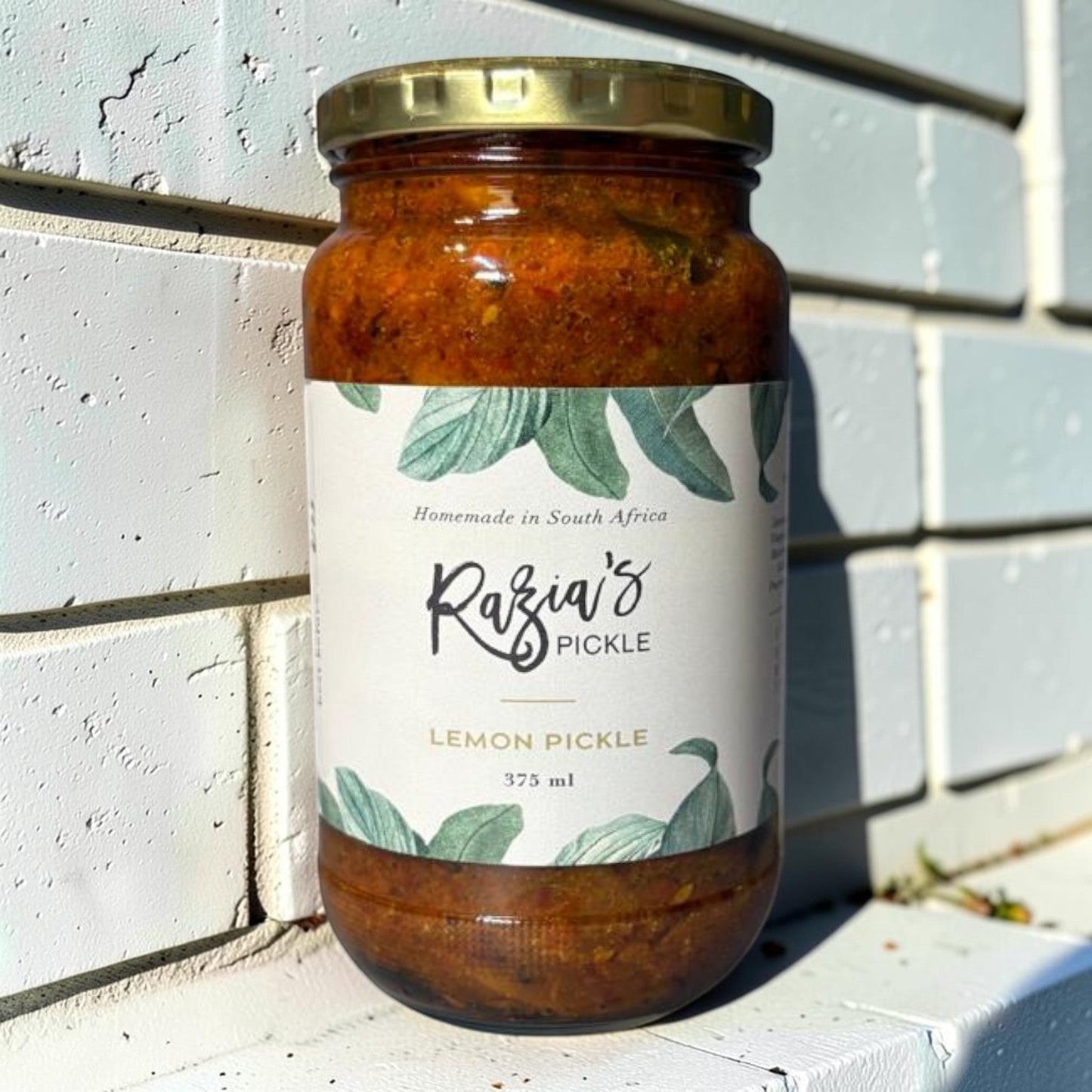 Die Spens Clarens Razia's Lemon Pickle in a glass jar with a gold lid, featuring a label with green leaves and text. Homemade in South Africa.