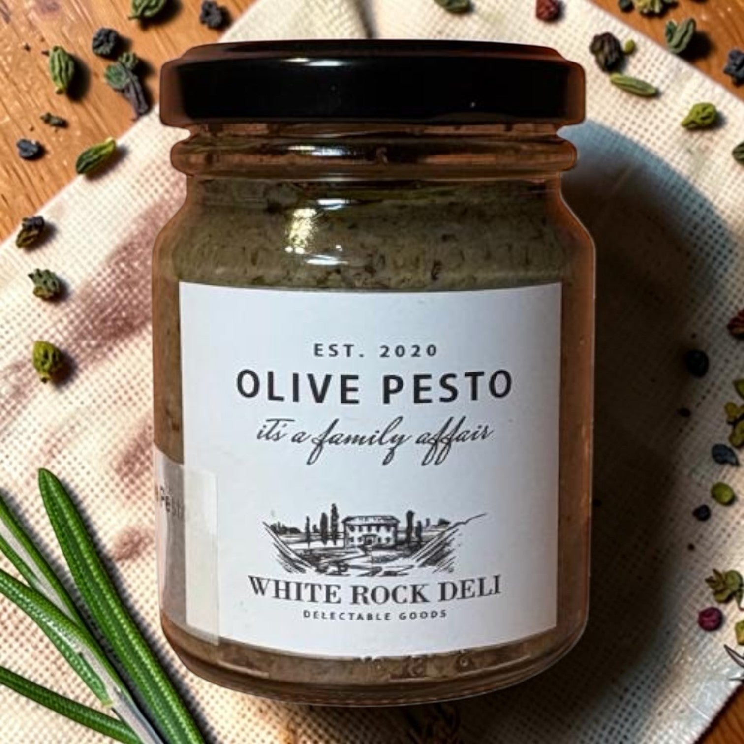 Die Spens Clarens White Rock Deli Olive Pesto in a glass jar with a black lid, garnished with rosemary and peppercorns.