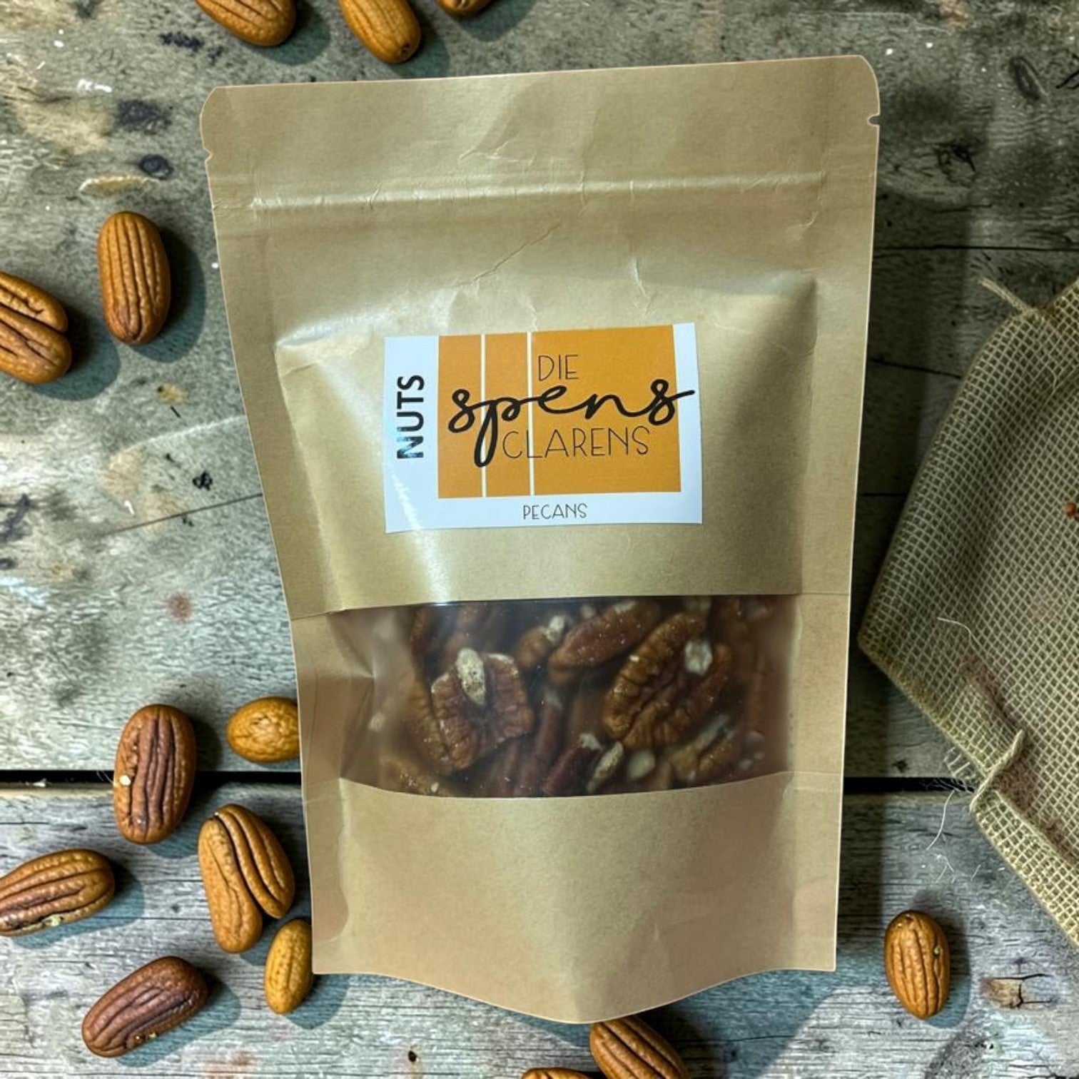 Die Spens Clarens resealable kraft bag filled with whole, rich pecan nuts. Scattered pecans on a wooden surface.