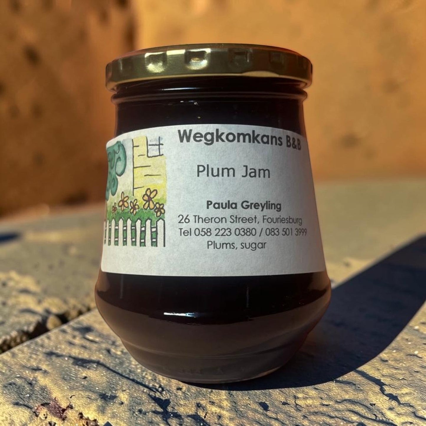 Die Spens Clarens Wegkomkans B&B plum jam in a glass jar with a gold lid. The label is white with a drawing of a fence and flowers, and text detailing the product and contact information.