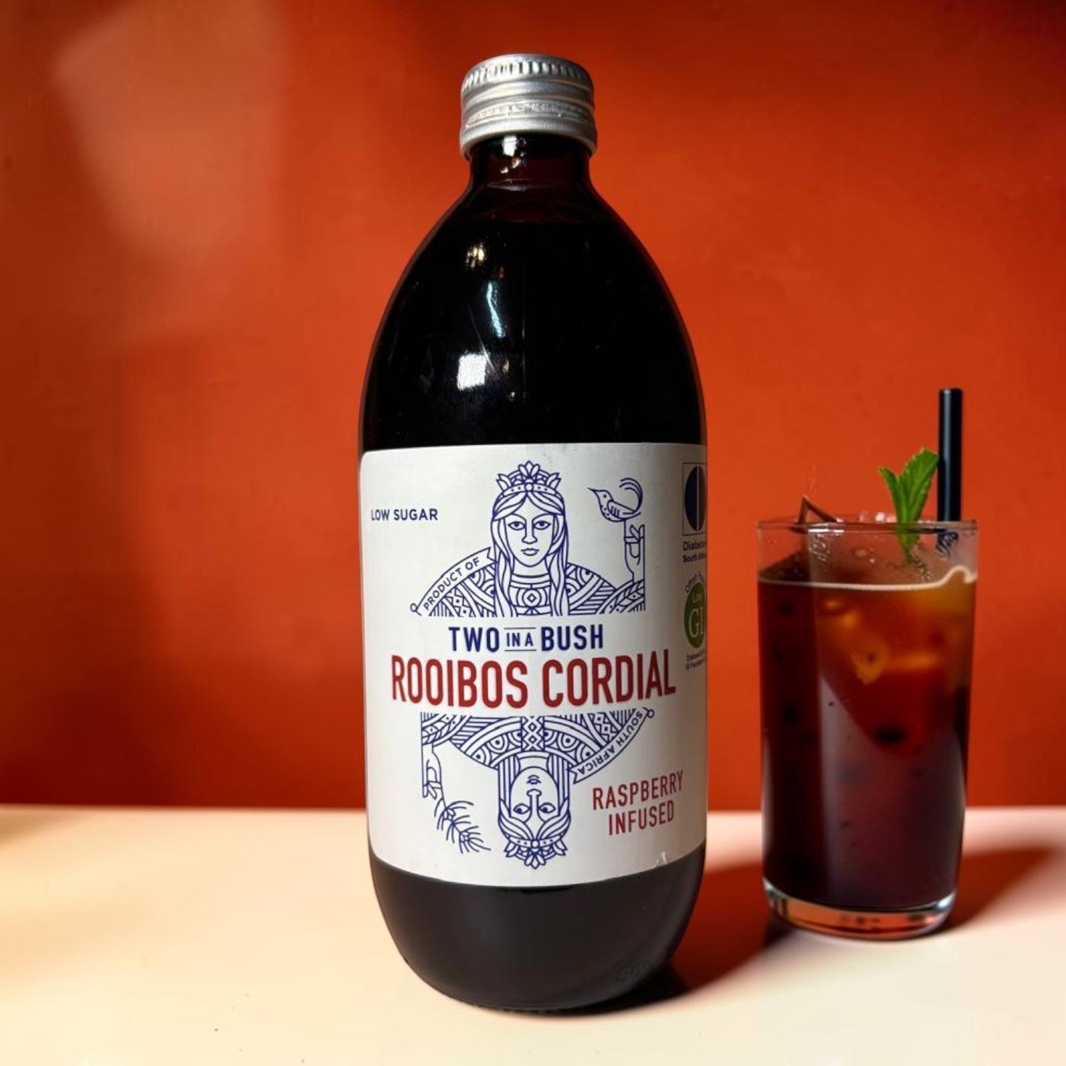 Die Spens Clarens Two In A Bush Raspberry Infused Rooibos Cordial bottle next to a glass of iced cordial.