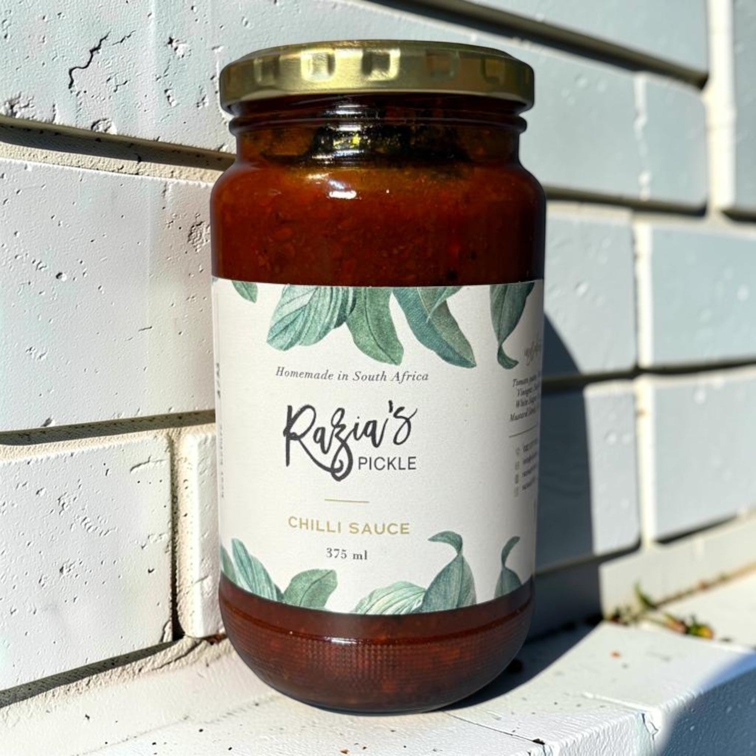 Die Spens Clarens Razia's Chilli Sauce, a homemade South African condiment in a glass jar with a gold lid. Features a white label with green leaf illustrations and text.