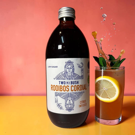 Die Spens Clarens Two In A Bush low sugar citrus infused rooibos cordial bottle next to a glass with a refreshing drink, lemon slice, and mint.