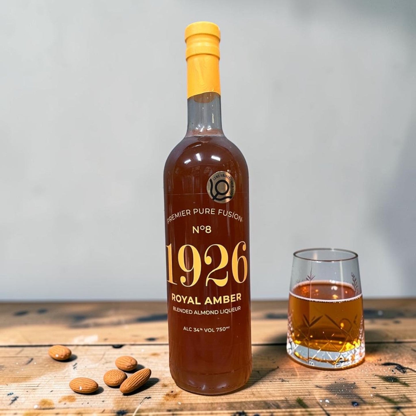 Die Spens Clarens Premier Pure Fusion 1926 Royal Amber blended almond liqueur in a glass bottle with a glass of liqueur and almonds on a wooden table.
