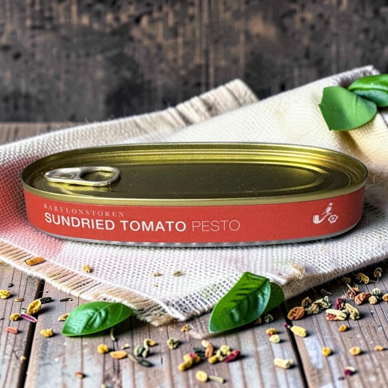 Die Spens Clarens Babylonstoren Sundried Tomato Pesto in a red and gold tin, garnished with basil leaves and scattered spices.