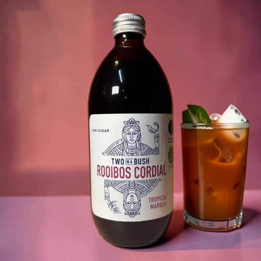 Die Spens Clarens Two In A Bush Tropical Marula low sugar rooibos cordial bottle next to a glass of iced drink.
