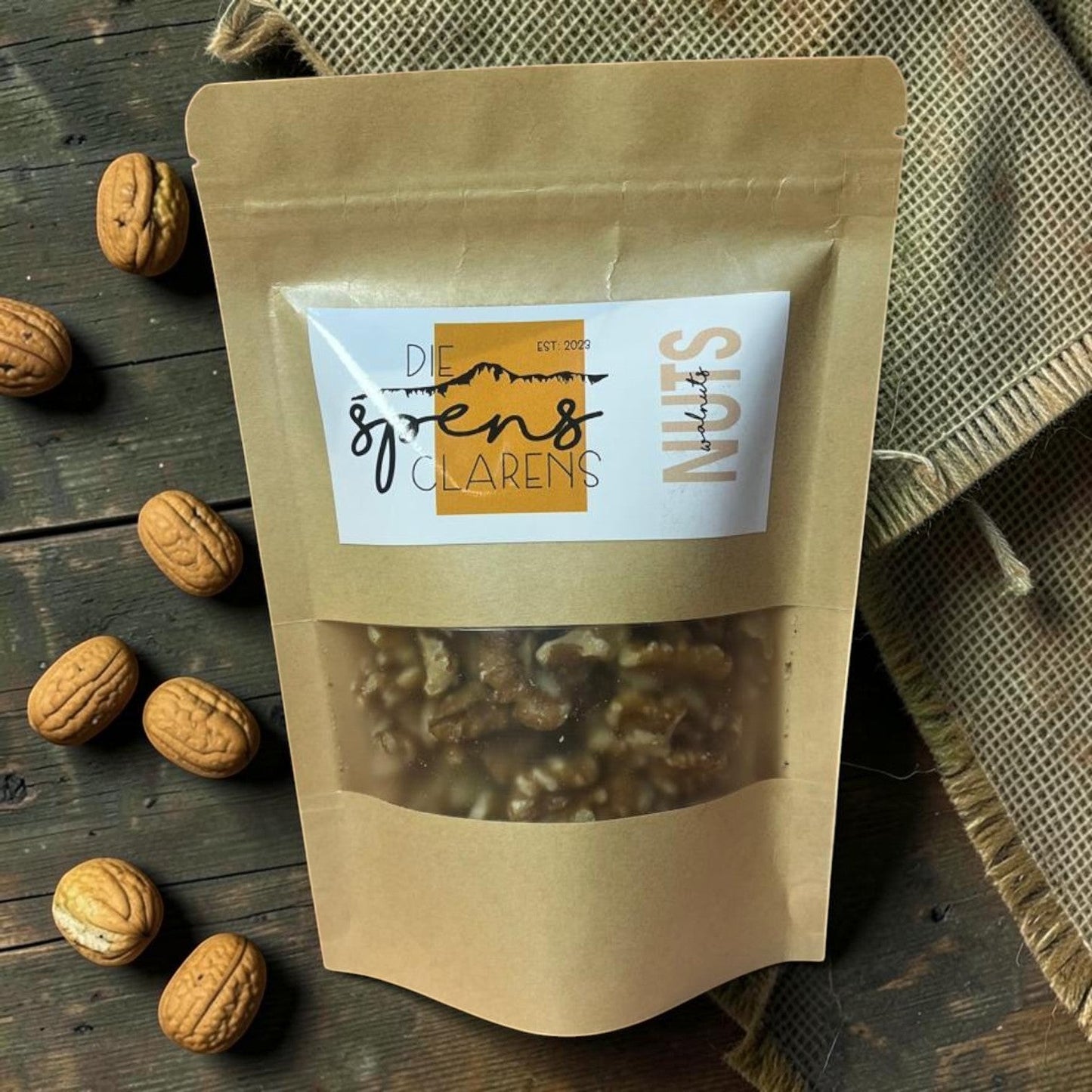 Die Spens Clarens premium quality walnuts in a resealable brown paper pouch, displayed on a rustic wooden surface with loose walnuts scattered around.