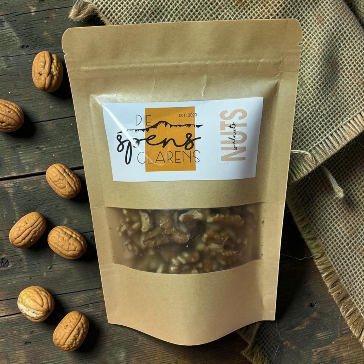 Die Spens Clarens premium quality walnuts in a resealable brown paper pouch, displayed on a rustic wooden surface with loose walnuts scattered around.