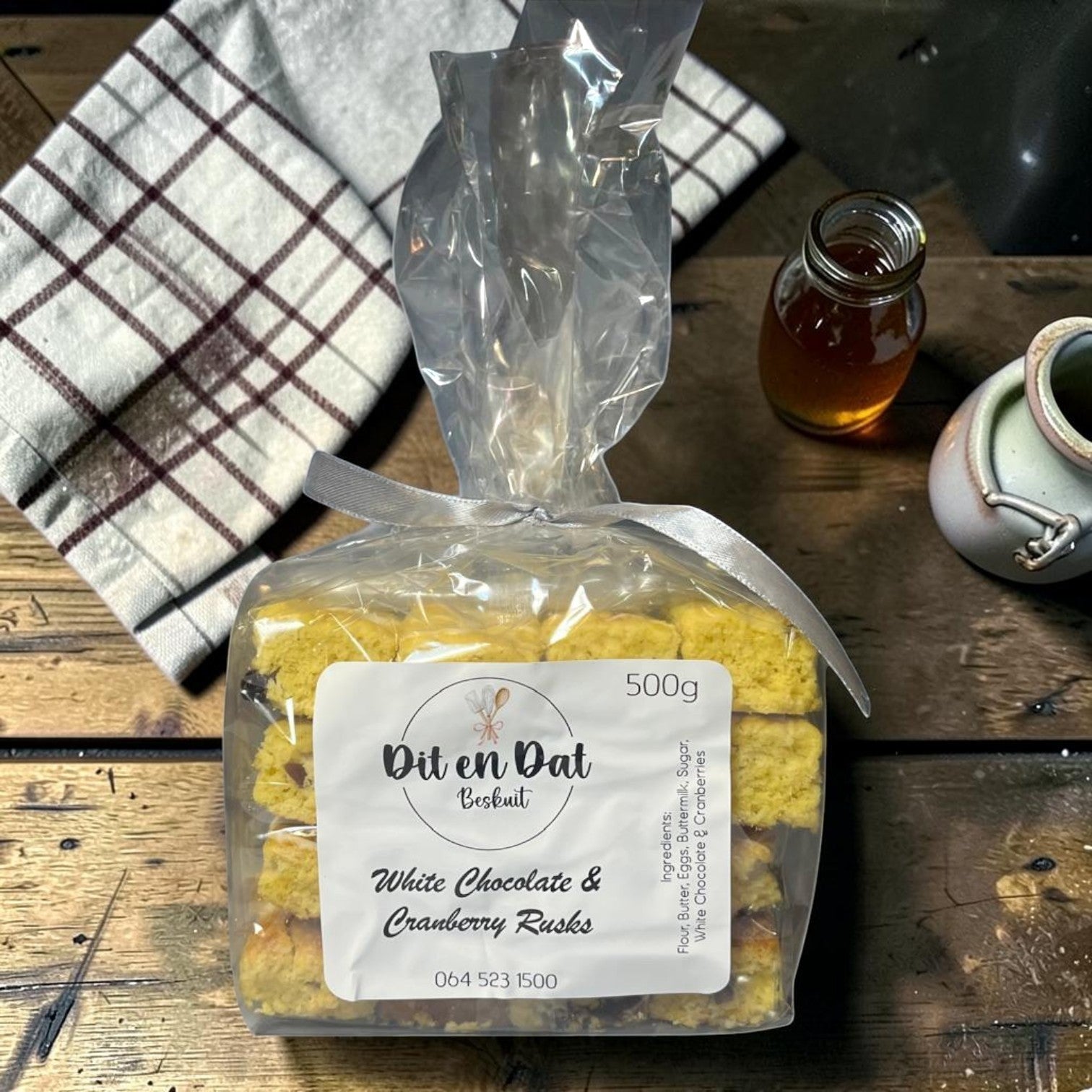 Die Spens Clarens White Chocolate & Cranberry Rusks in a clear bag, 500g pack, with a checkered cloth and honey.
