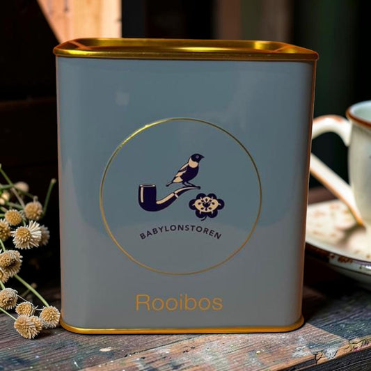 Die Spens Clarens grey tin with gold trim, featuring a circular emblem with a bird on a pipe and the text Babylonstoren. Rooibos tea is written in gold lettering.