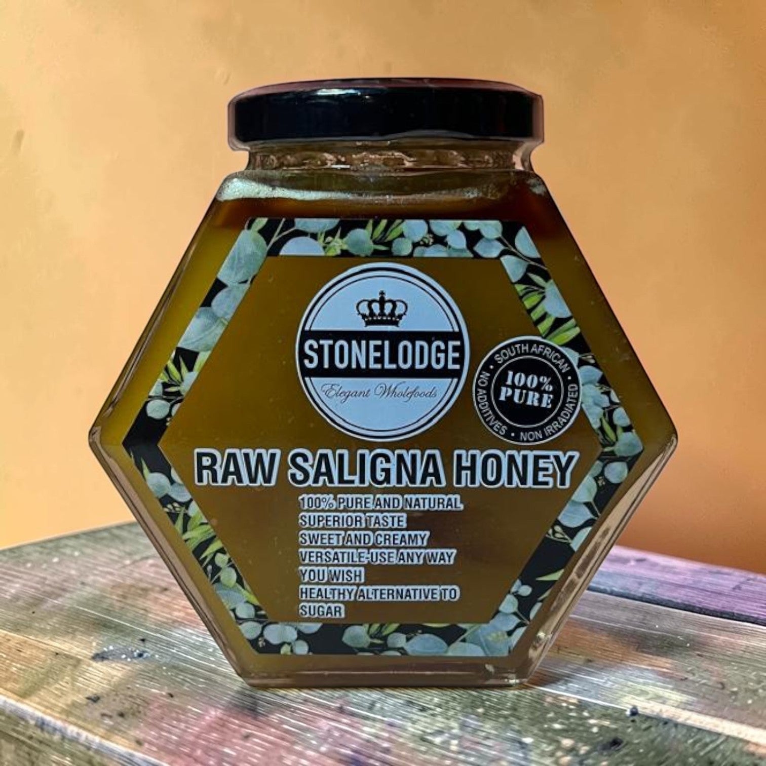 Die Spens Clarens Stonelodge Raw Saligna Honey in a hexagonal glass jar with a black lid. The label is golden with floral accents and text detailing its pure, natural, sweet, and creamy qualities.